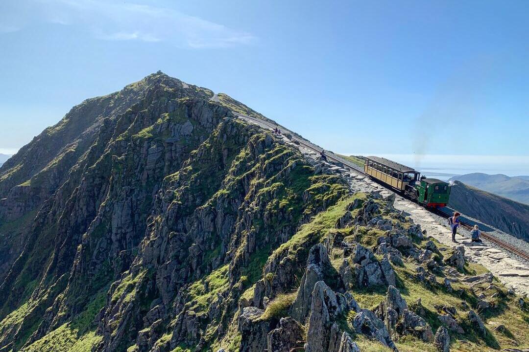 Mountain landscape with a steep railway, a train ascending, rocky ridges, greenery, and clear blue sky. People walk beside the track.