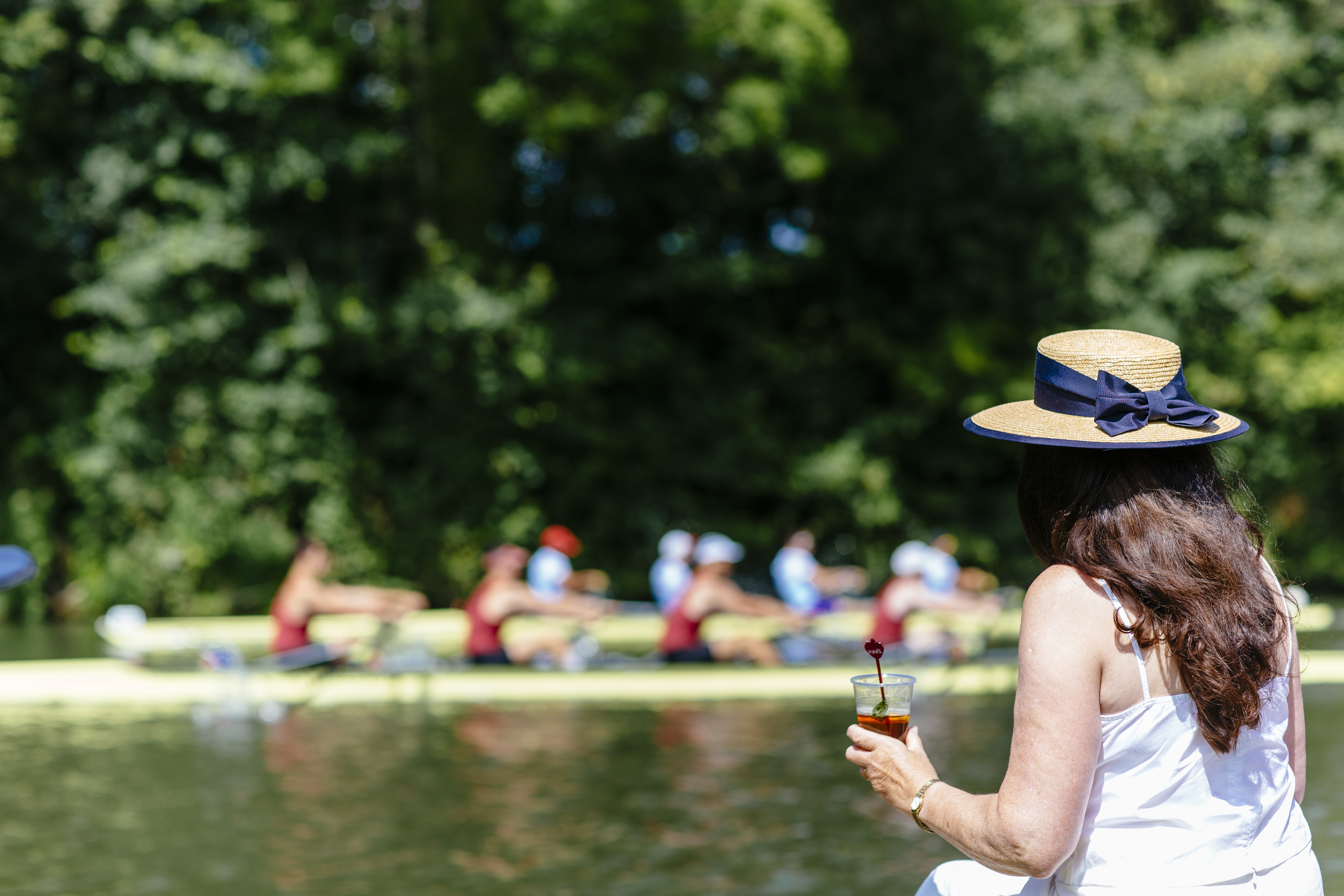 Une femme portant un chapeau, buvant tout en regardant une course de bateaux