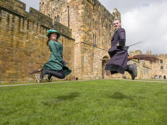 Two wizarding professors demonstrating flying on a broom outside a large castle.