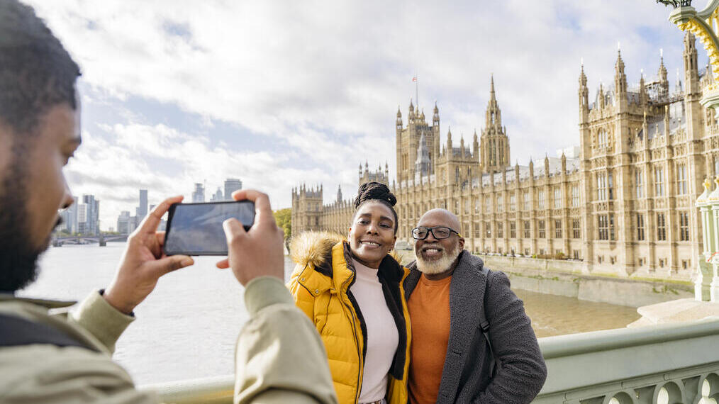 A man photographing mature couple with a large river and iconic buildings in the background.