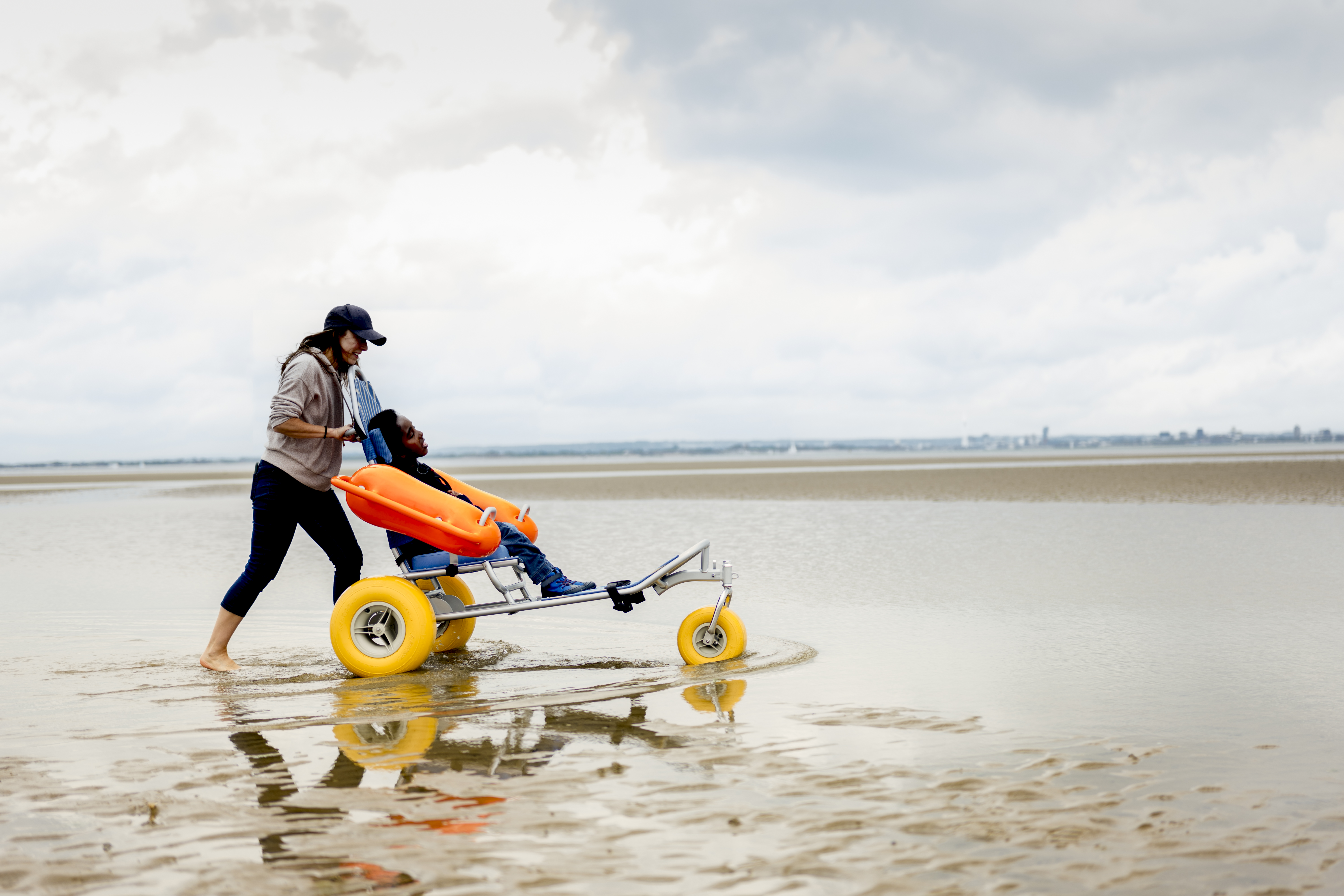 Eine Frau mit ihrem Sohn, der in einem barrierefreien Meeresrollstuhl mit Schwimmkörpern am Strand sitzt.