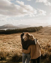 Zwei Freunde umarmen sich und blicken über das Tal auf einen See und die Berge