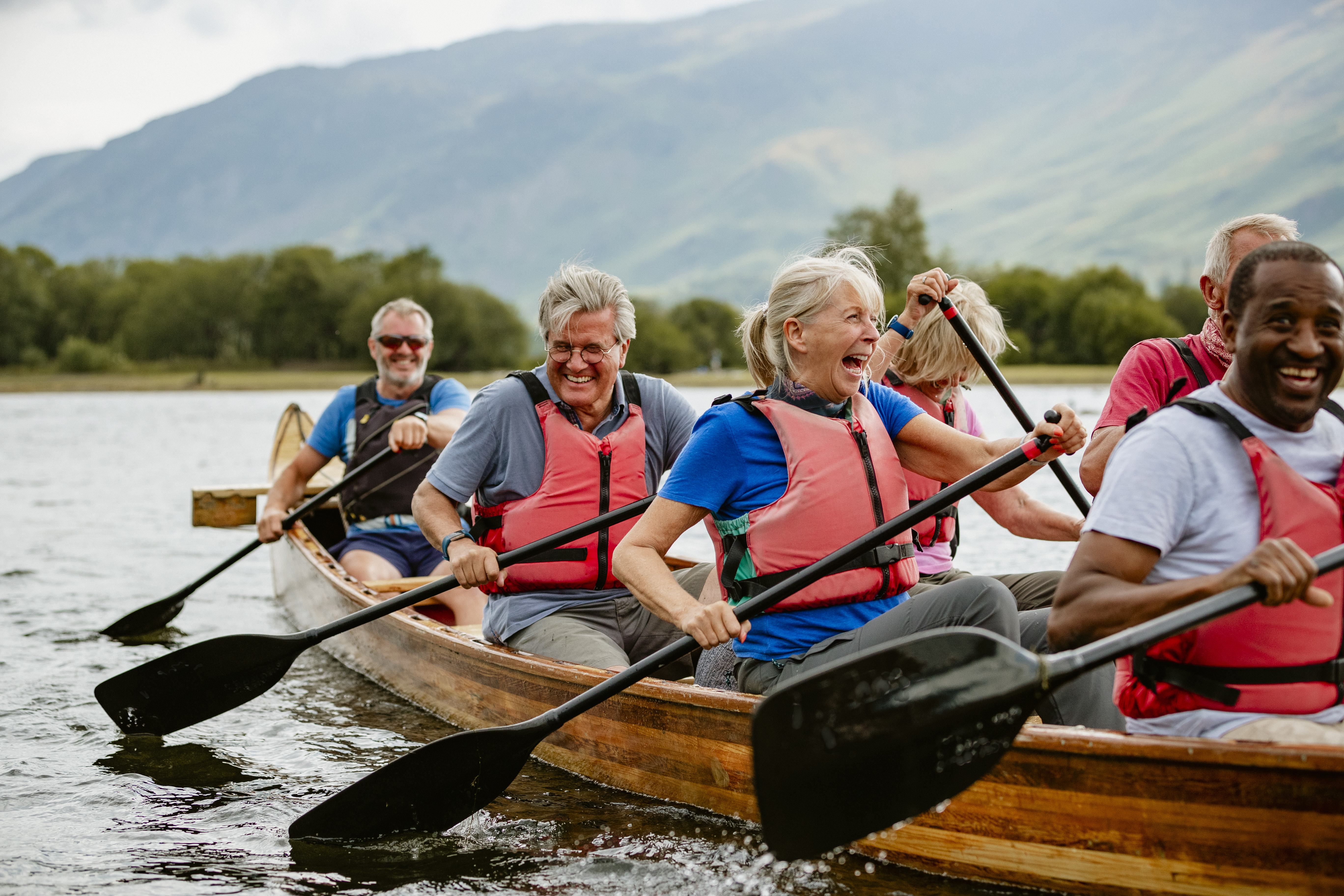 A senior group of friends wearing lifejackets enjoying rowing in a single boat on a river.