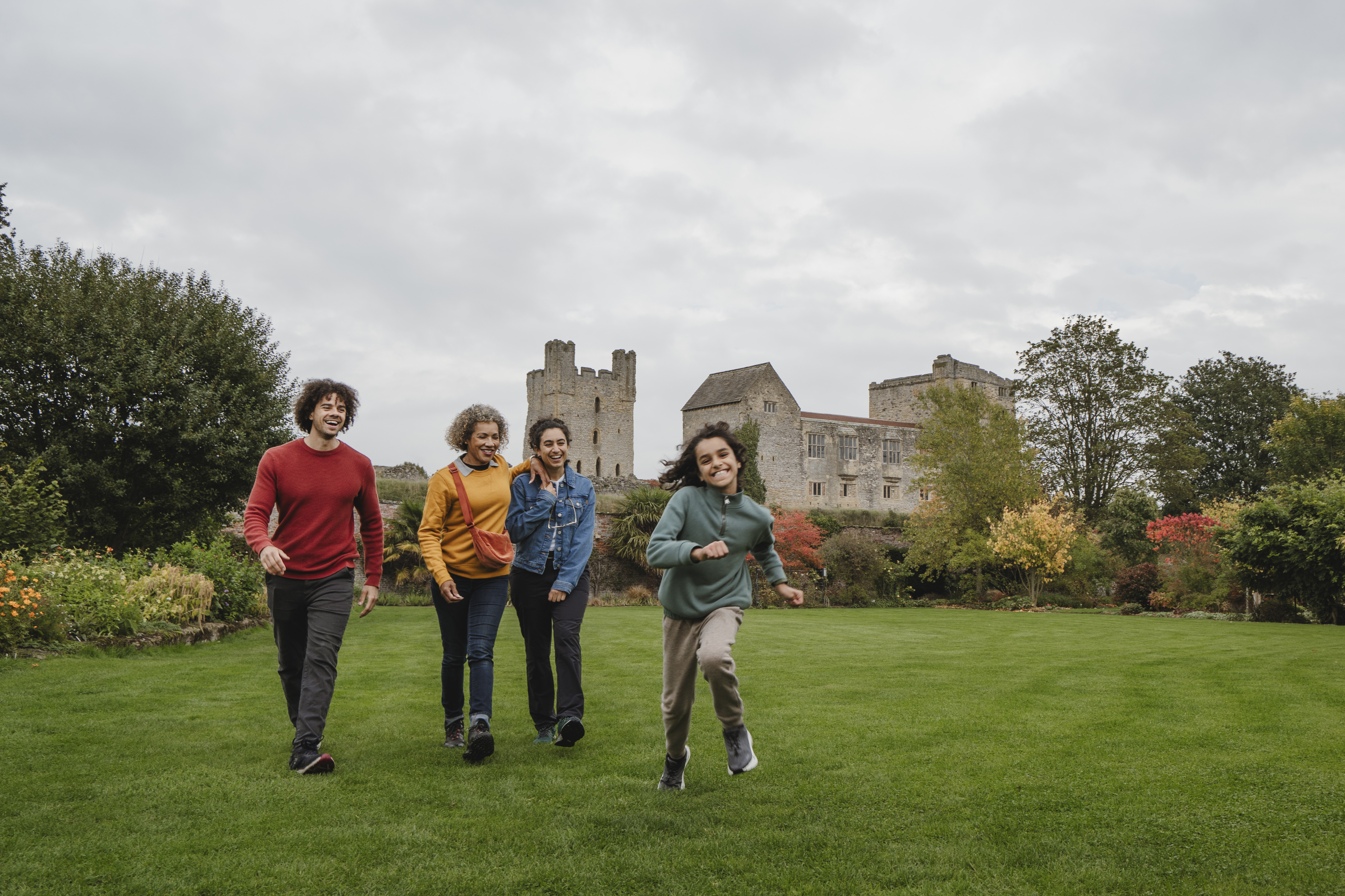 Four people walking on a large green lawn with an old stone castle and autumn trees in the background, under a cloudy sky.