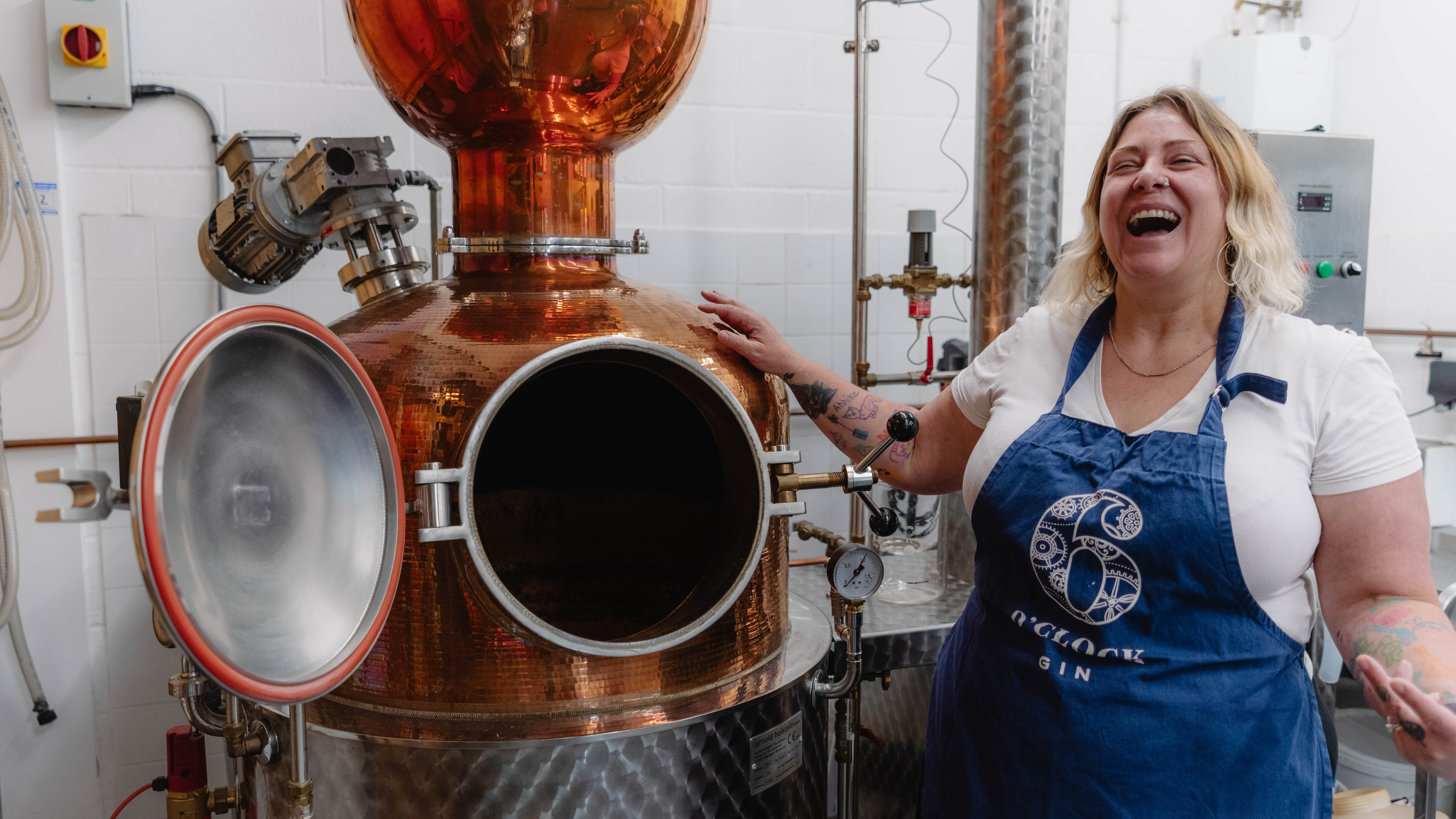 A female tour guide wears an apron and stands by a still at a distillery