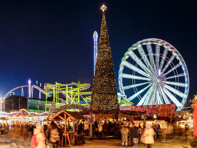 A Christmas fair at night with a large decorated Christmas tree, ferris wheel, roller coaster, lights, and crowds of people.