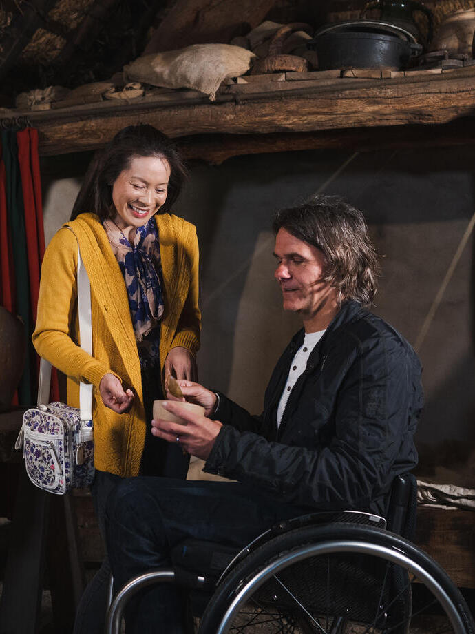 Man and woman, inside a recreated 15th century cottage looking at domestic utensils, man in a wheelchair