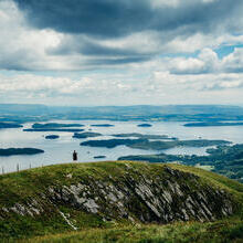 A wide panorama of a hiker standing atop a hill looking out over grassland and lakes.