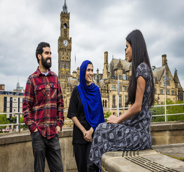 Two women and a man stand and talk in front of a town view