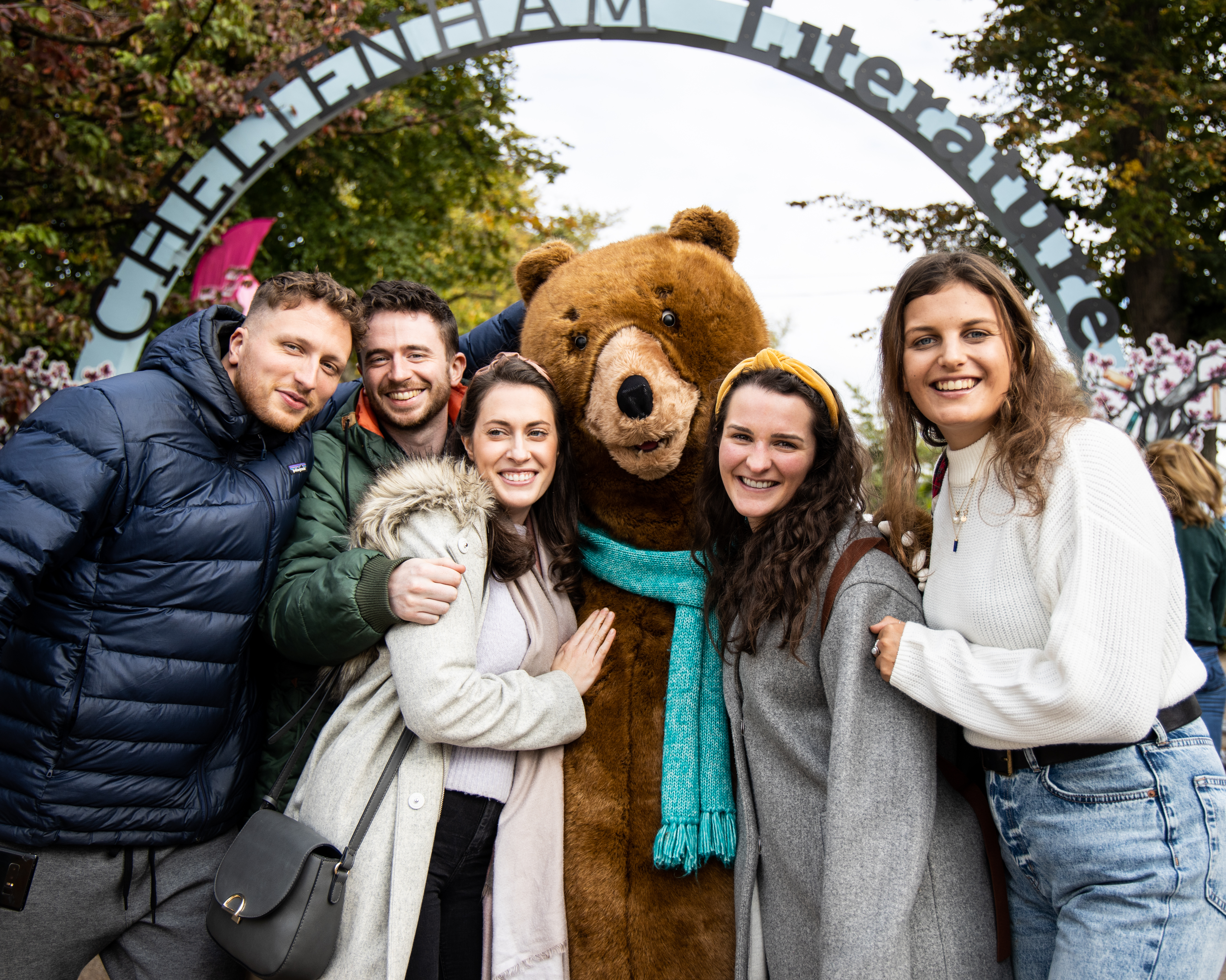 People posing with big bear mascot under an arch sign reading Cheltenham Literature
