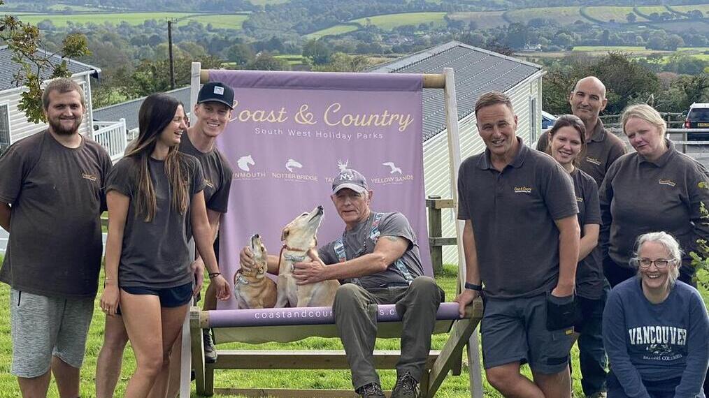 Group of people and a dog posing by a Coast & Country sign outdoors, with rolling countryside and lodges in the background.