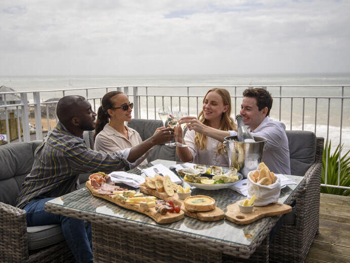 Two men and two women laugh together while sat on a hotel balcony with the sea beyond