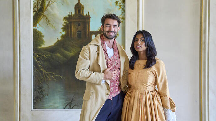 Ein Mann und eine Frau in historischen Kostümen stehen in einem dekorierten Raum vor einem Gemälde mit Landschaft und altem Gebäude.