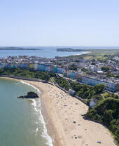 An overhead shot of a sandy beach lined with sunbathers, surrounded by brightly coloured townhouses and countryside.
