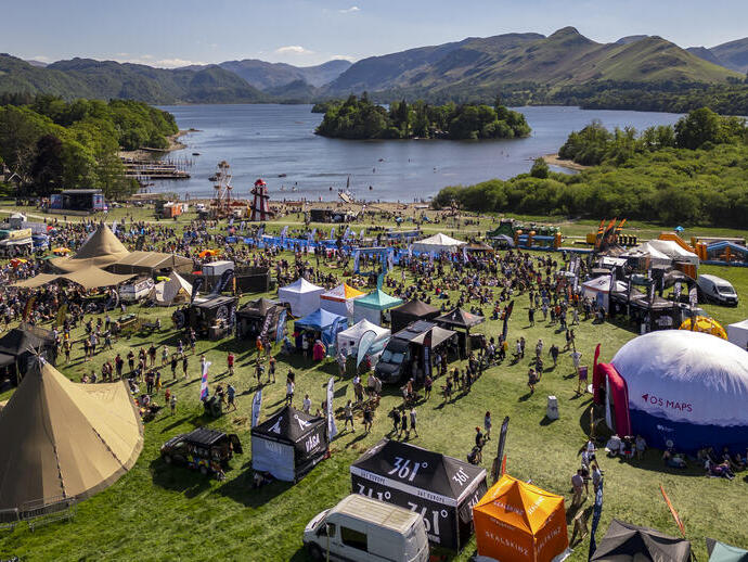 Outdoor festival with tents, food trucks, and crowds by a lake, surrounded by mountains and lush greenery on a sunny day.