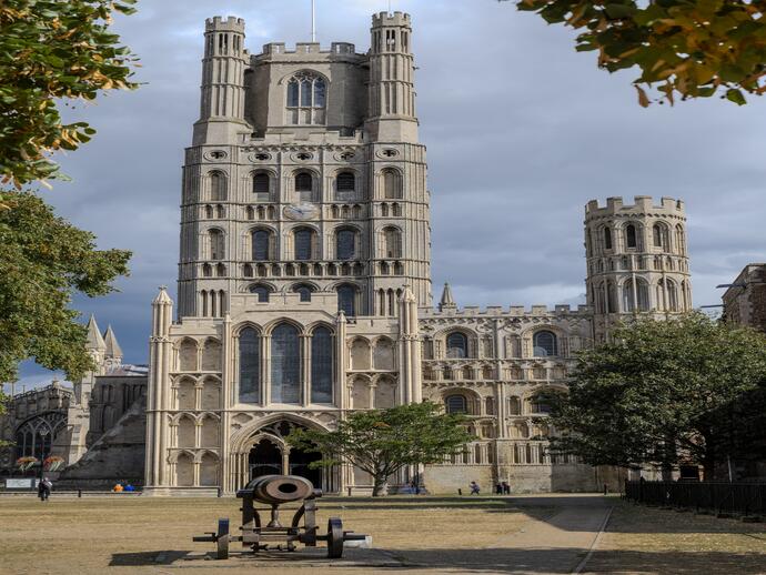 Large historic cathedral with intricate architecture, tall towers, and a cannon in the foreground, surrounded by trees and cloudy sky.