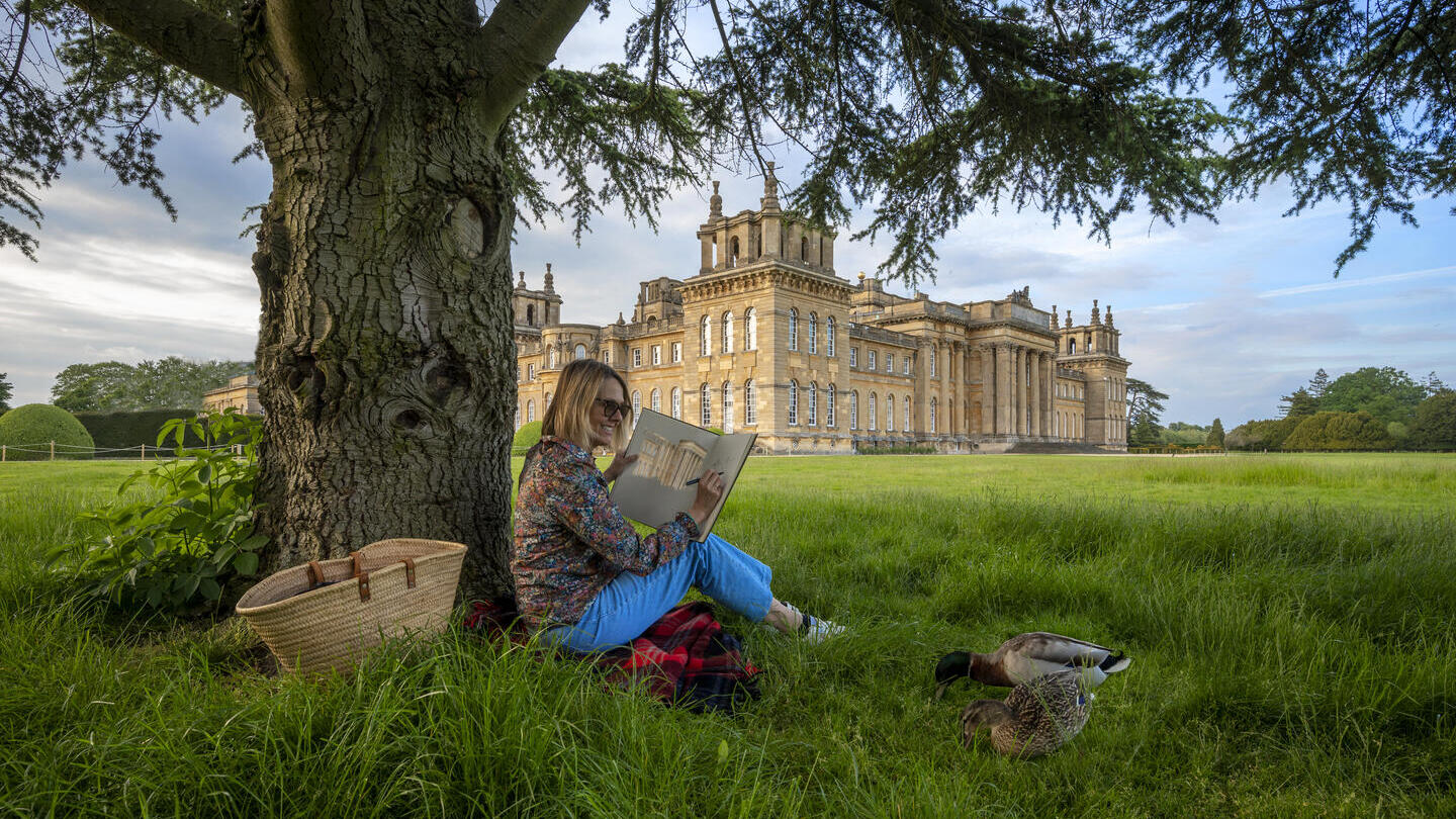 Woman sat under a tree sketching in the grounds of a large country estate
