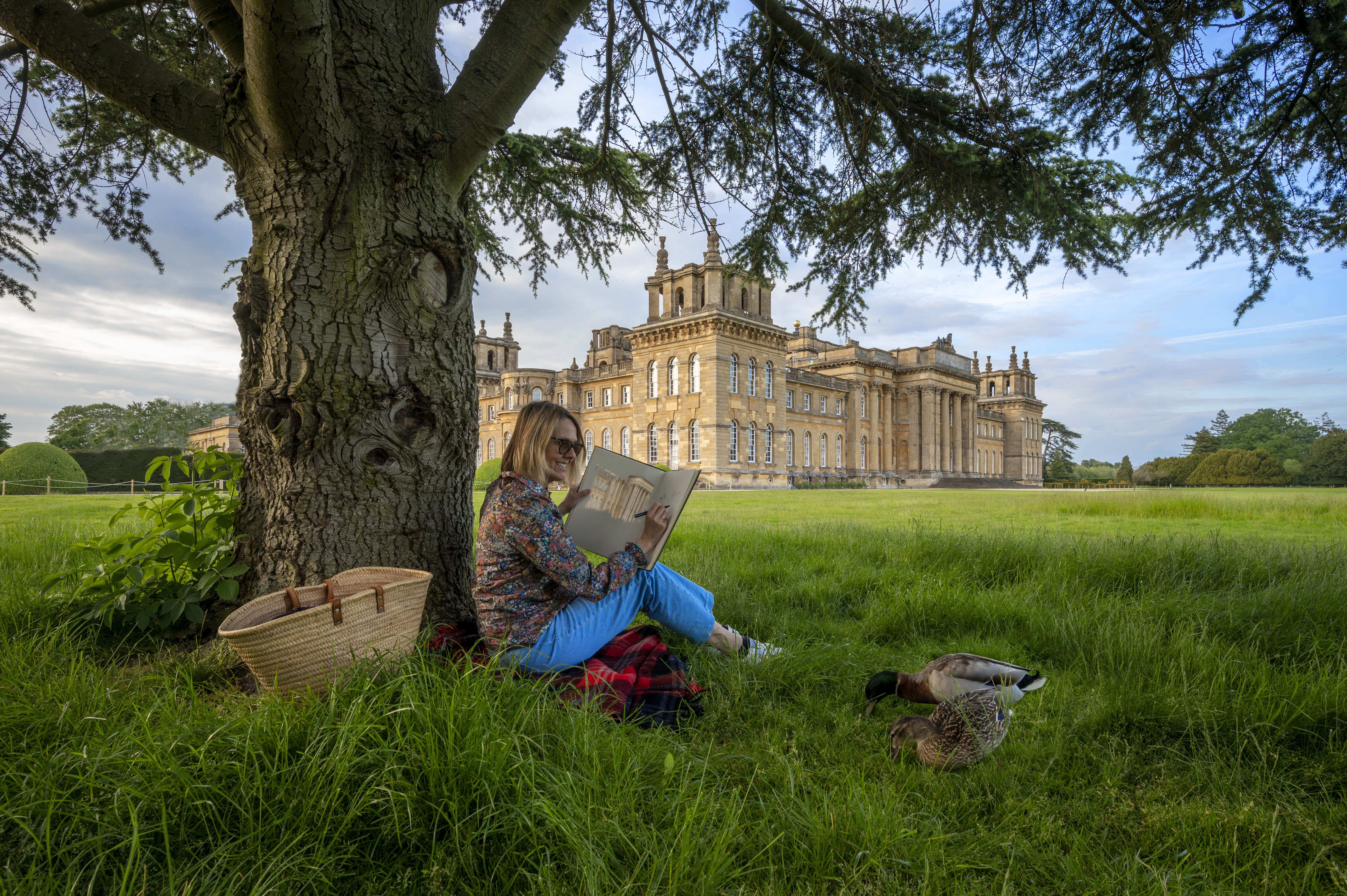 Une femme assise sous un arbre en train de dessiner dans le parc d'un grand domaine rural