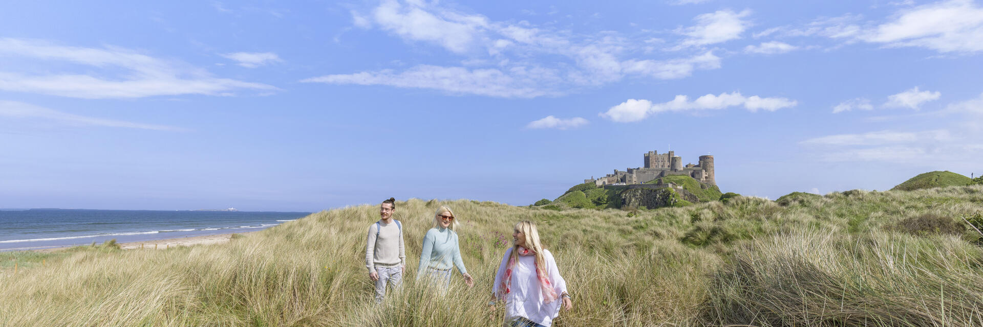 A group of people walk among tall grasses in dunes with a castle in the background