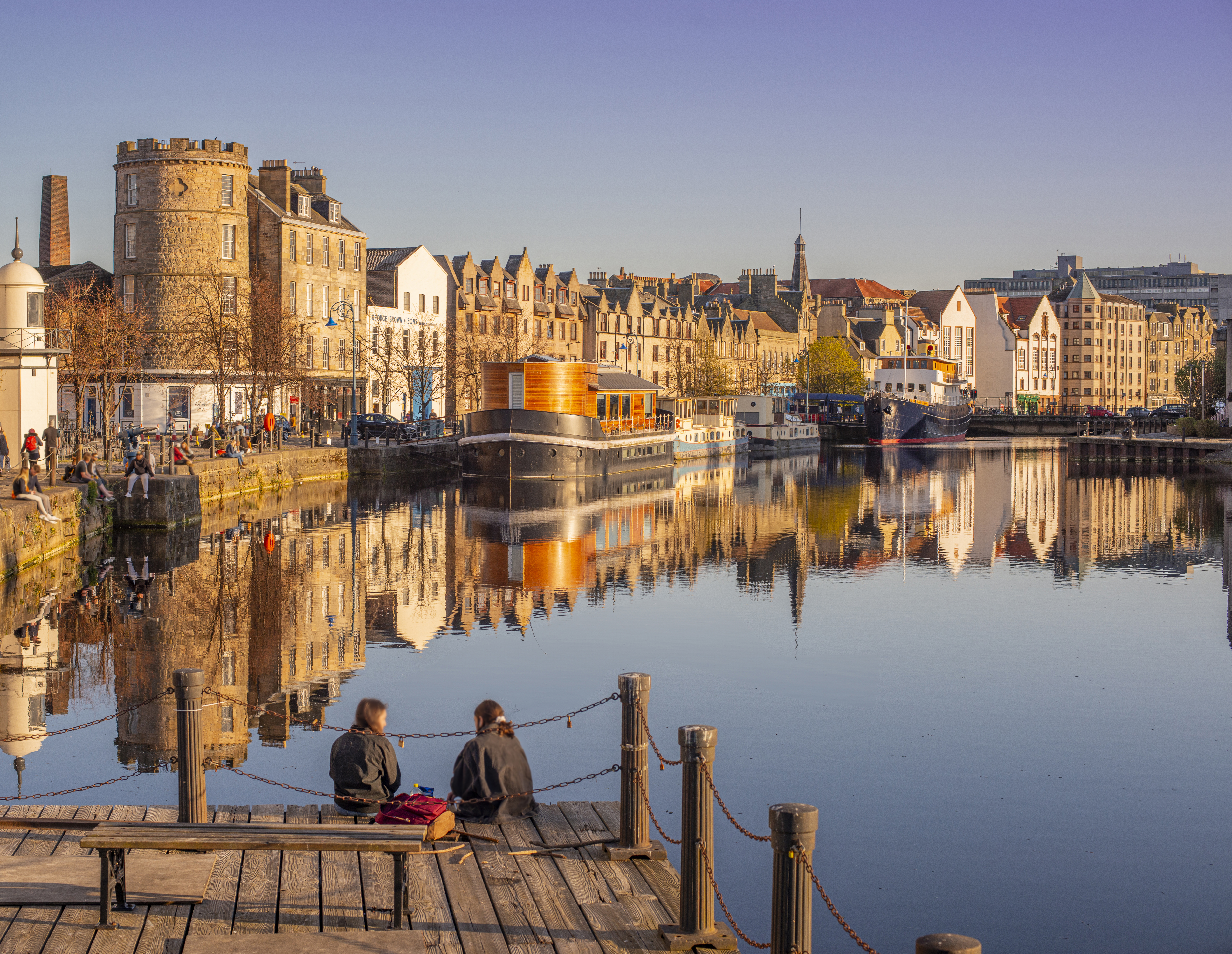 Dos jóvenes sentadas en el embarcadero del paseo marítimo al atardecer, con barcos y edificios junto al puerto.