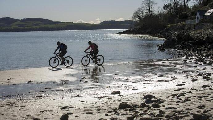 Two cyclists ride along a rocky shoreline at low tide, with calm water, distant hills, trees, and a house visible in the background.