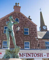 Bronze statue of a person blowing a horn in front of a red stone building and clock tower on a bright, clear day.