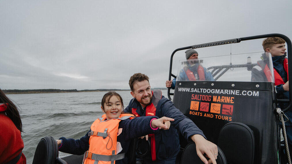 A man and a girl point at something from inside a boat with the Marine boat crew in the background.