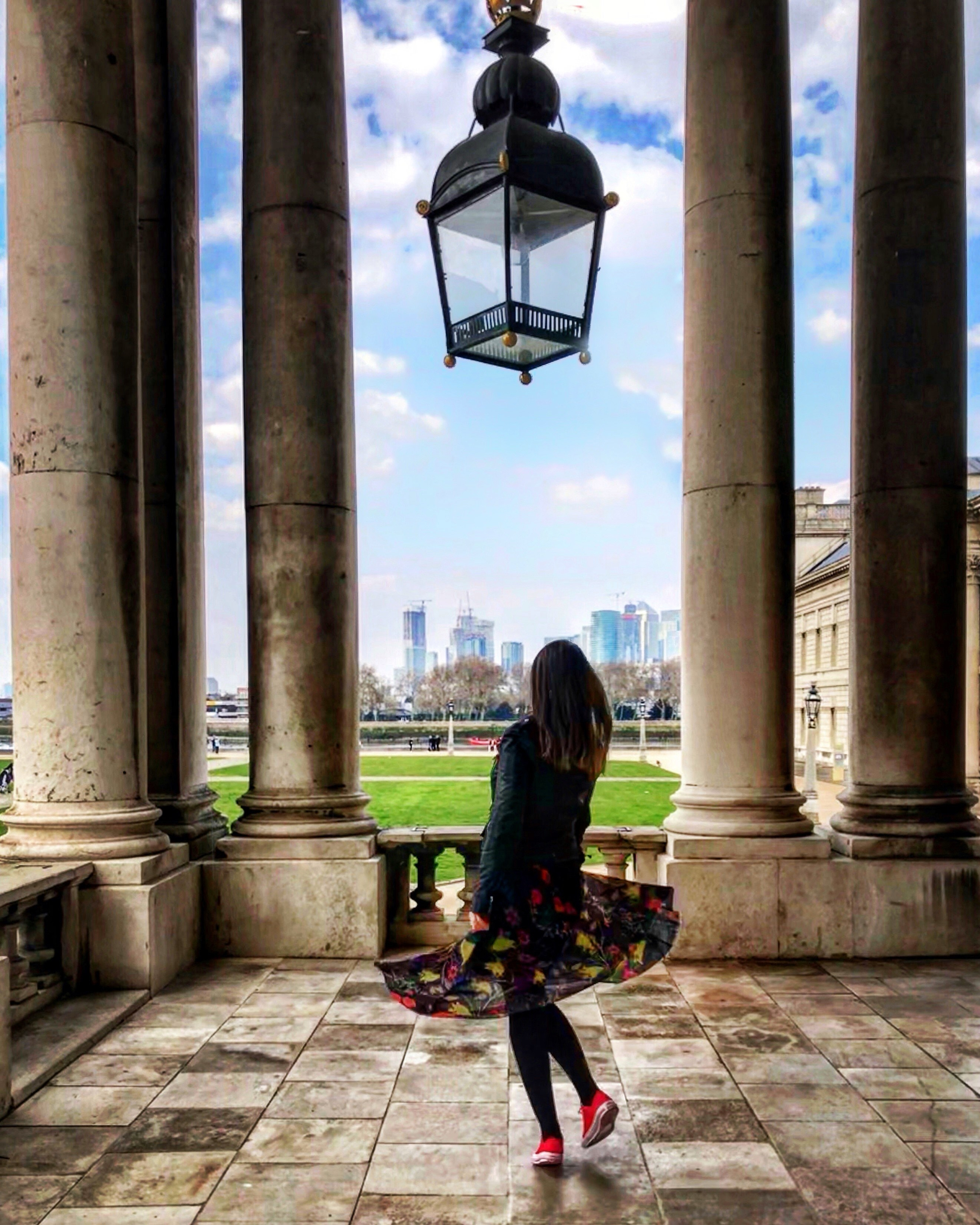 Woman standing underneath a large glass lantern, looking through the colonnades towards modern city buildings