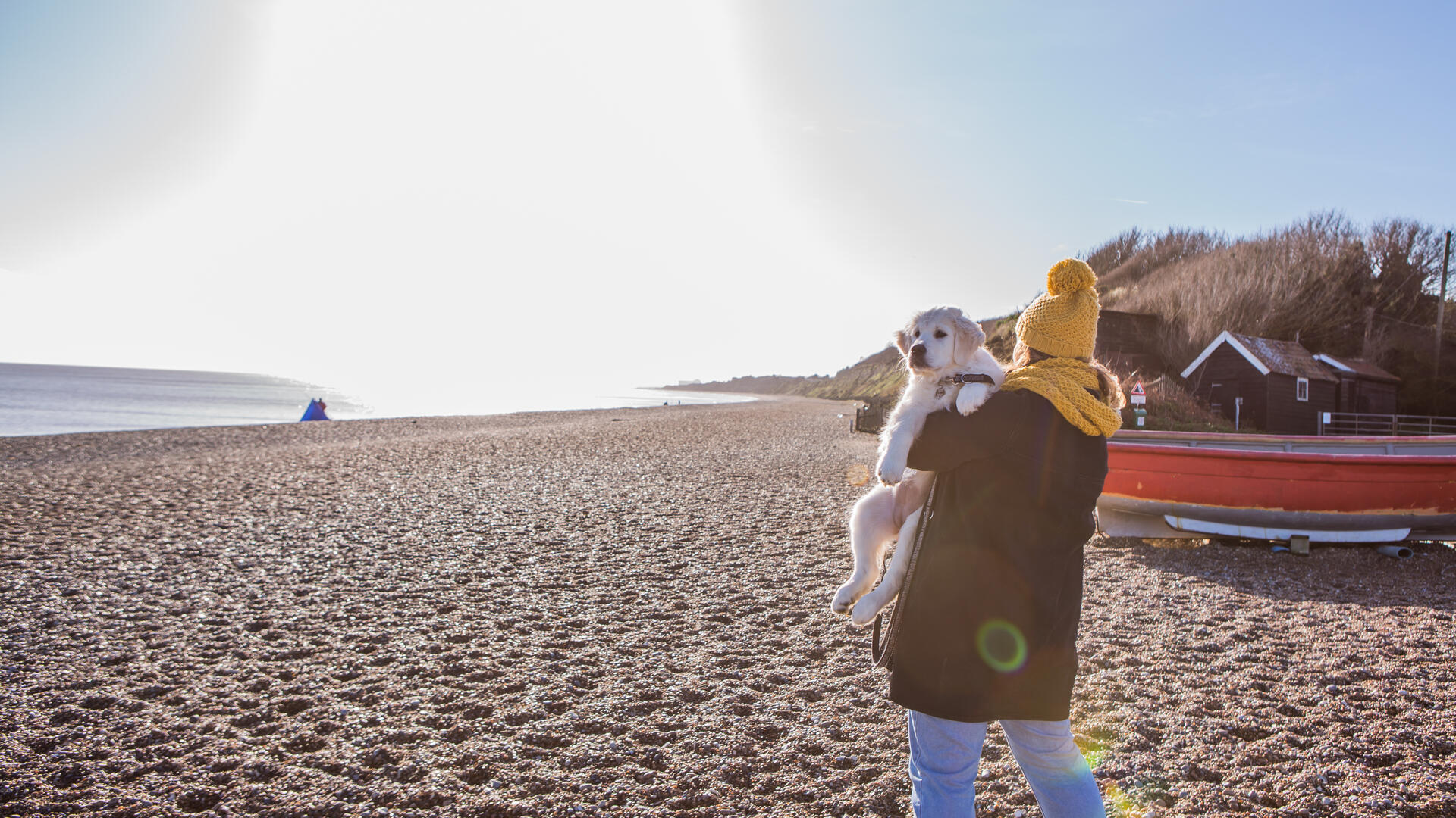 A woman carrying a dog along a beach in Dunwich