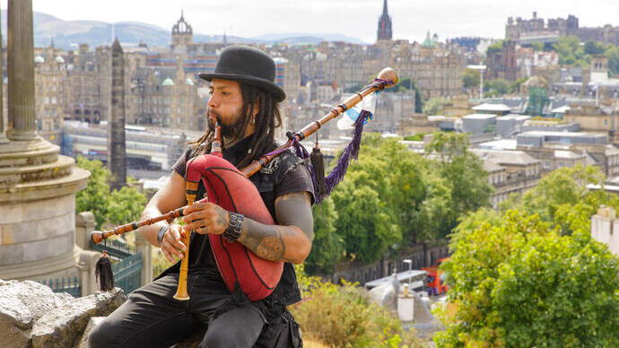 Man playing the bagpipes sat on a rock on a hill overlooking a city