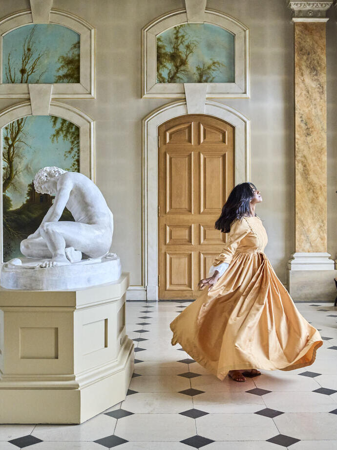 Mujer con vestido largo girando en un gran salón con escultura de mármol, columnas y paredes decoradas.