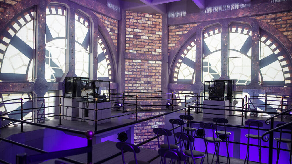 Interior with large clock face windows, exposed brick walls, metal railings, and rows of chairs lit by purple lighting.