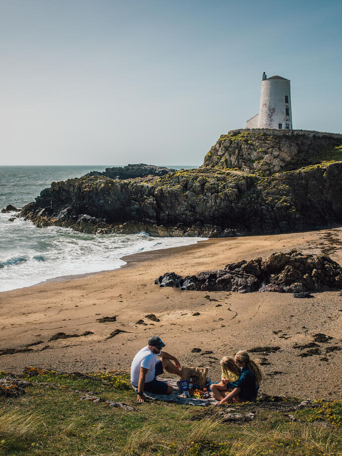Une jeune famille pique-niquant sur une plage avec un phare en arrière-plan.
