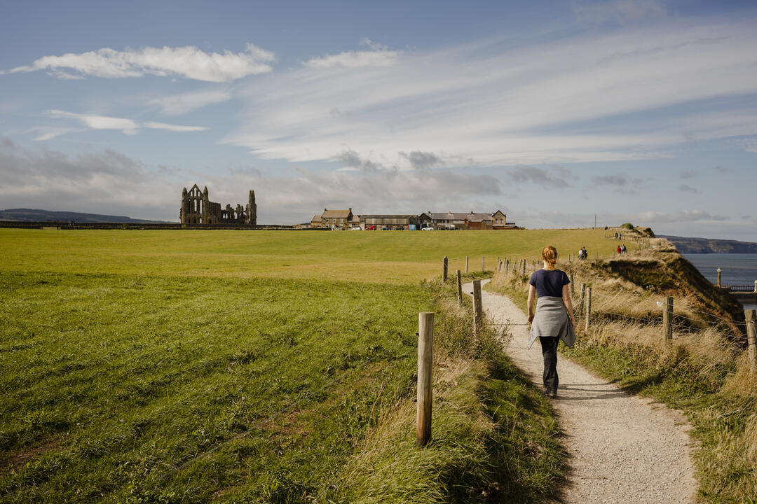 Woman walking along a coastal path towards a ruined abbey