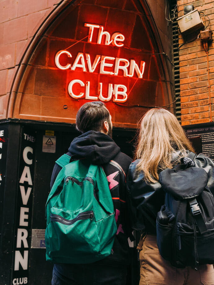 Two people with backpacks stand outside The Cavern Club, a famous brick music venue with neon signage in Liverpool.