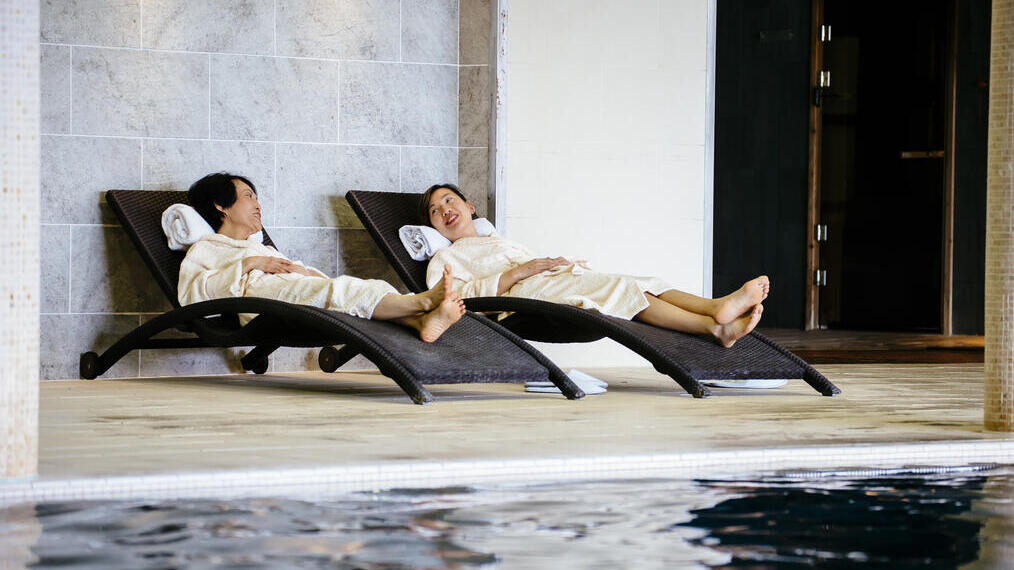 Two women lying on loungers beside an indoor swimming pool
