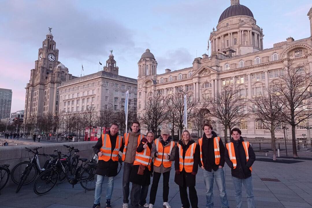 Group in orange safety vests standing by bicycles, in front of historic buildings at Liverpool waterfront at sunset.