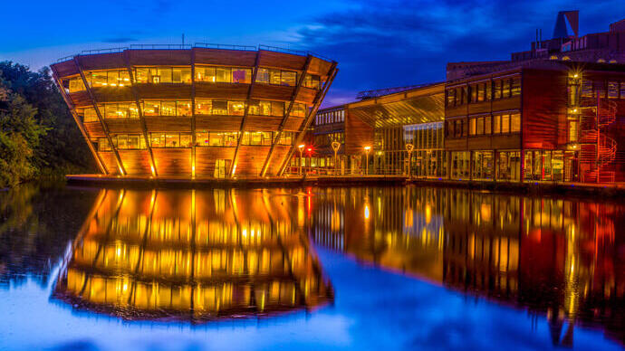 A modern, illuminated building with wooden panels is reflected in a calm body of water at night, against a deep blue sky.