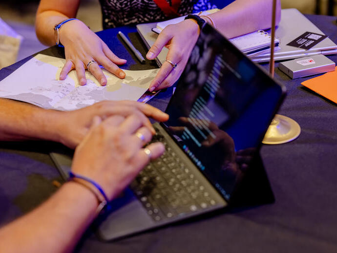 Two people work together at a table with a laptop, map, notebooks, and business cards, suggesting a collaborative or planning meeting.