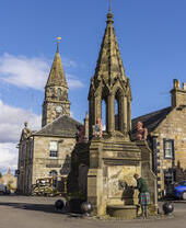 رجل يحصل على الماء من نافورة حجرية كبيرة مع تماثيل أسود حمراء باهتة في ساحة بلدة.