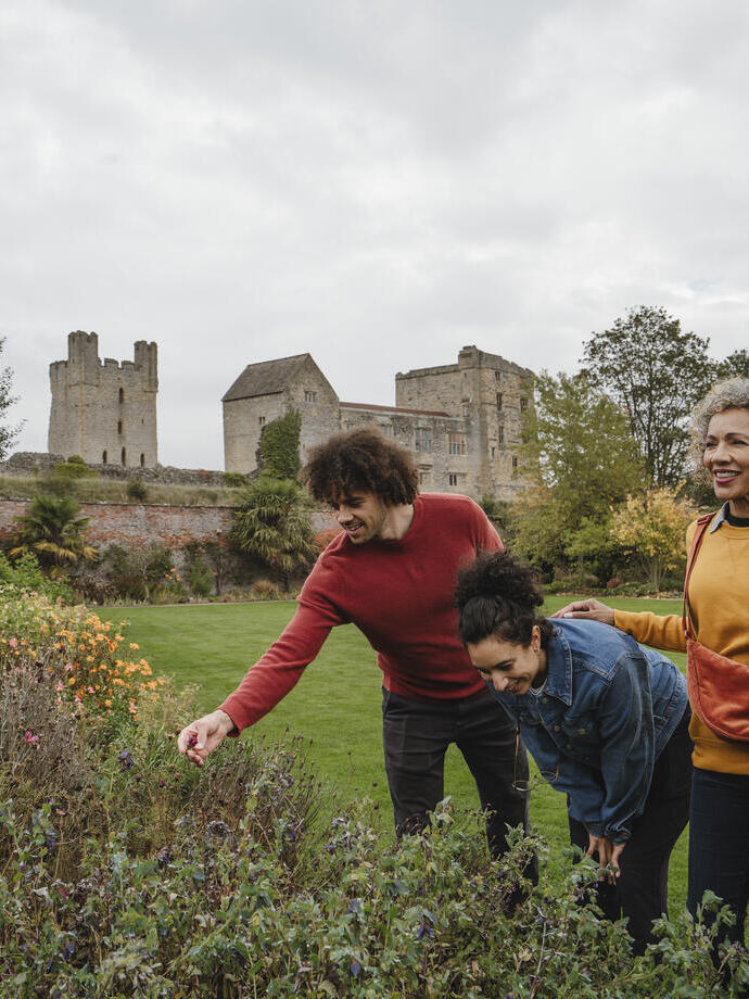 A group enjoys a flower garden with a historic stone castle and green lawns in the background on a cloudy day.
