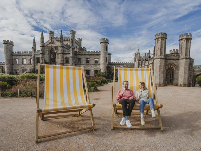 Un homme et une femme sont assis sur de grandes chaises de plage rayées jaunes et blanches devant un château historique, mangeant des glaces.
