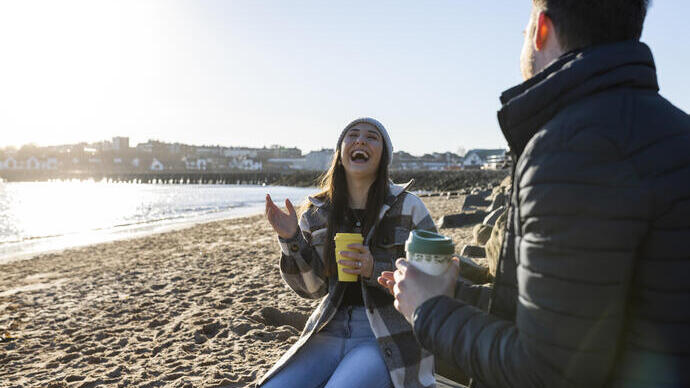 Two people sit on a sandy beach at sunset, laughing and holding takeaway coffee cups, with seaside buildings in the background.