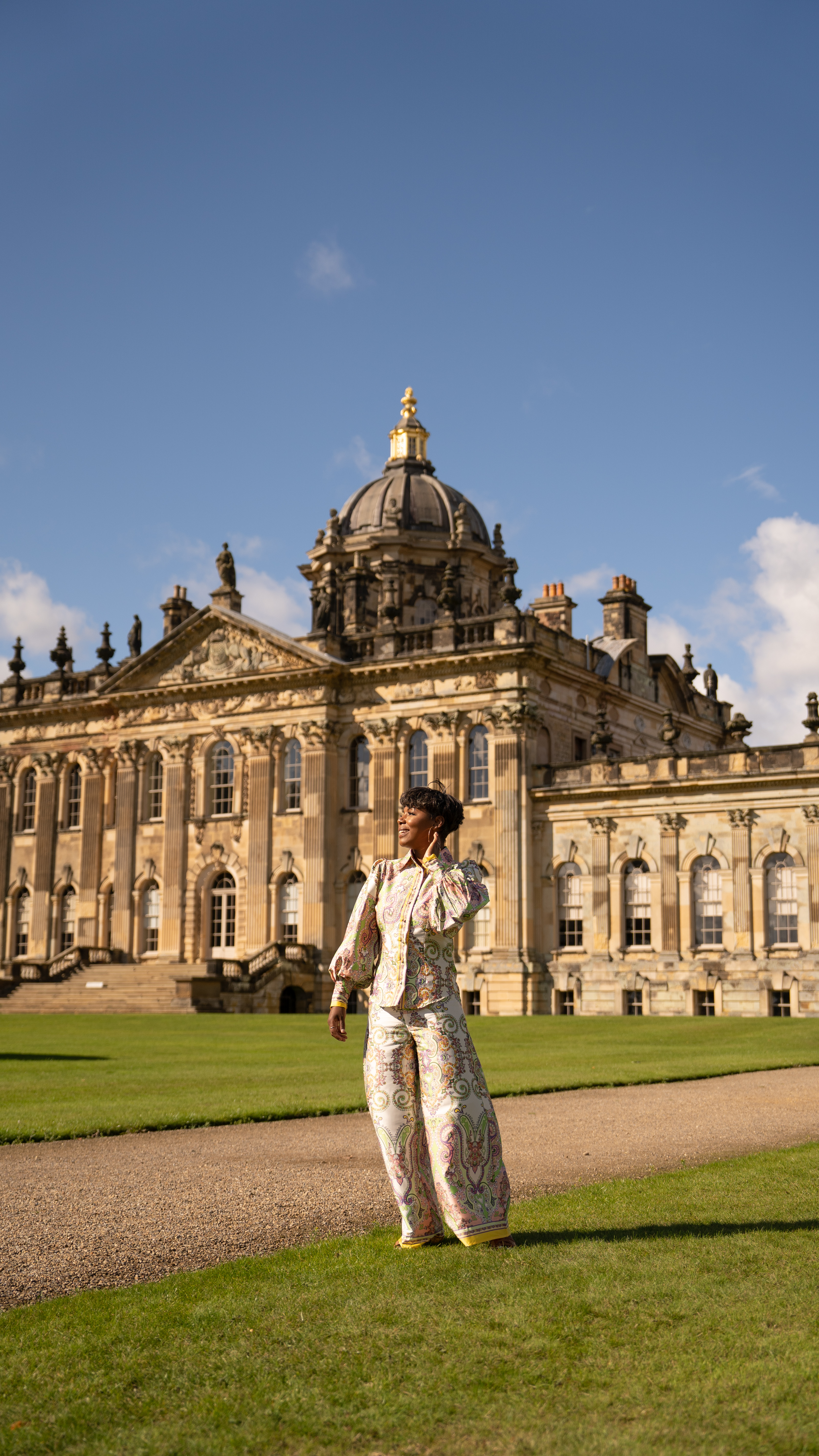 Person stands on a path in front of a grand historic building with ornate columns and a large domed roof under a blue sky.