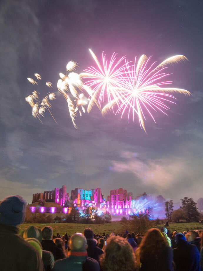 Fireworks display over a ruined castle watched by a crowd of people in the evening