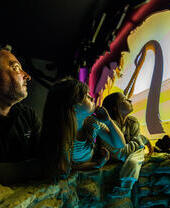 A man and two young girls watch an animated water monster with a long neck on a screen .