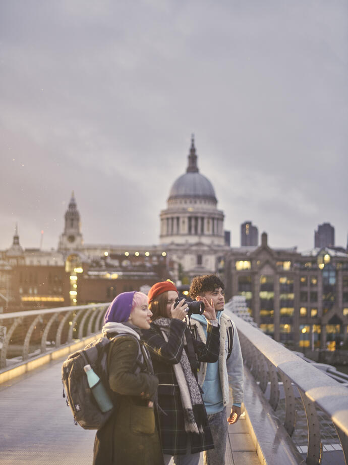 Two women and a man on a pedestrian bridge in a city taking photographs