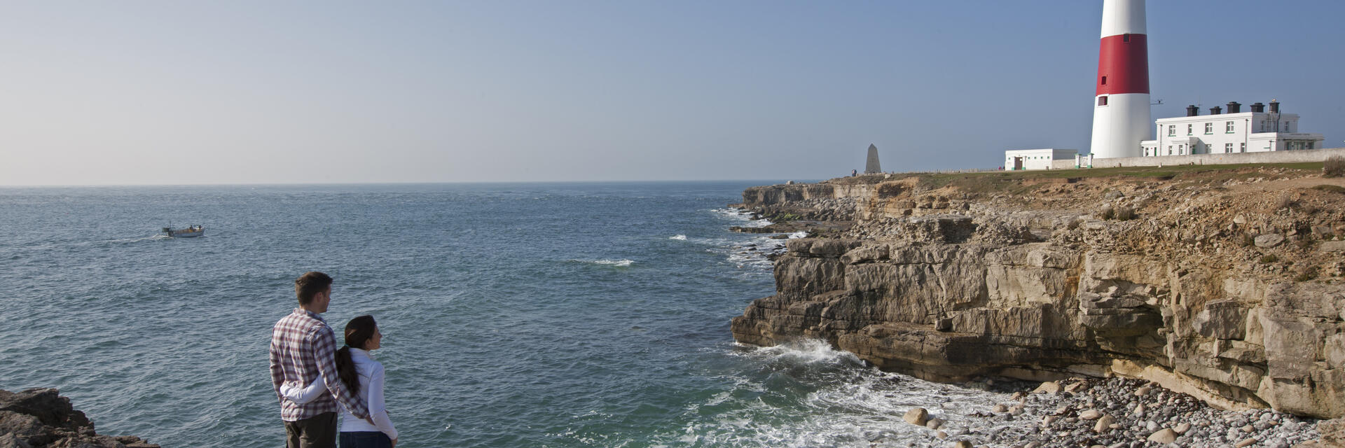 Couple standing on a cliff by the sea looking at a red and white lighthouse 