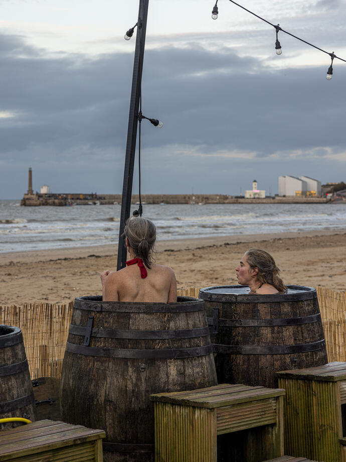 Two women in barrels in an outdoor beachside sauna.
