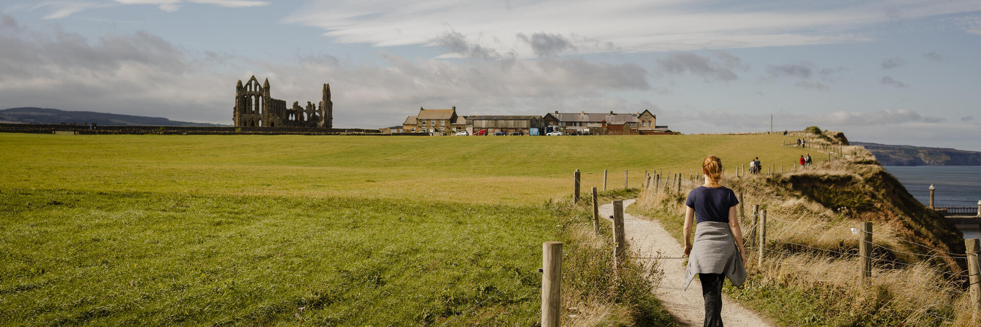 Woman walking along a coastal path towards a ruined abbey